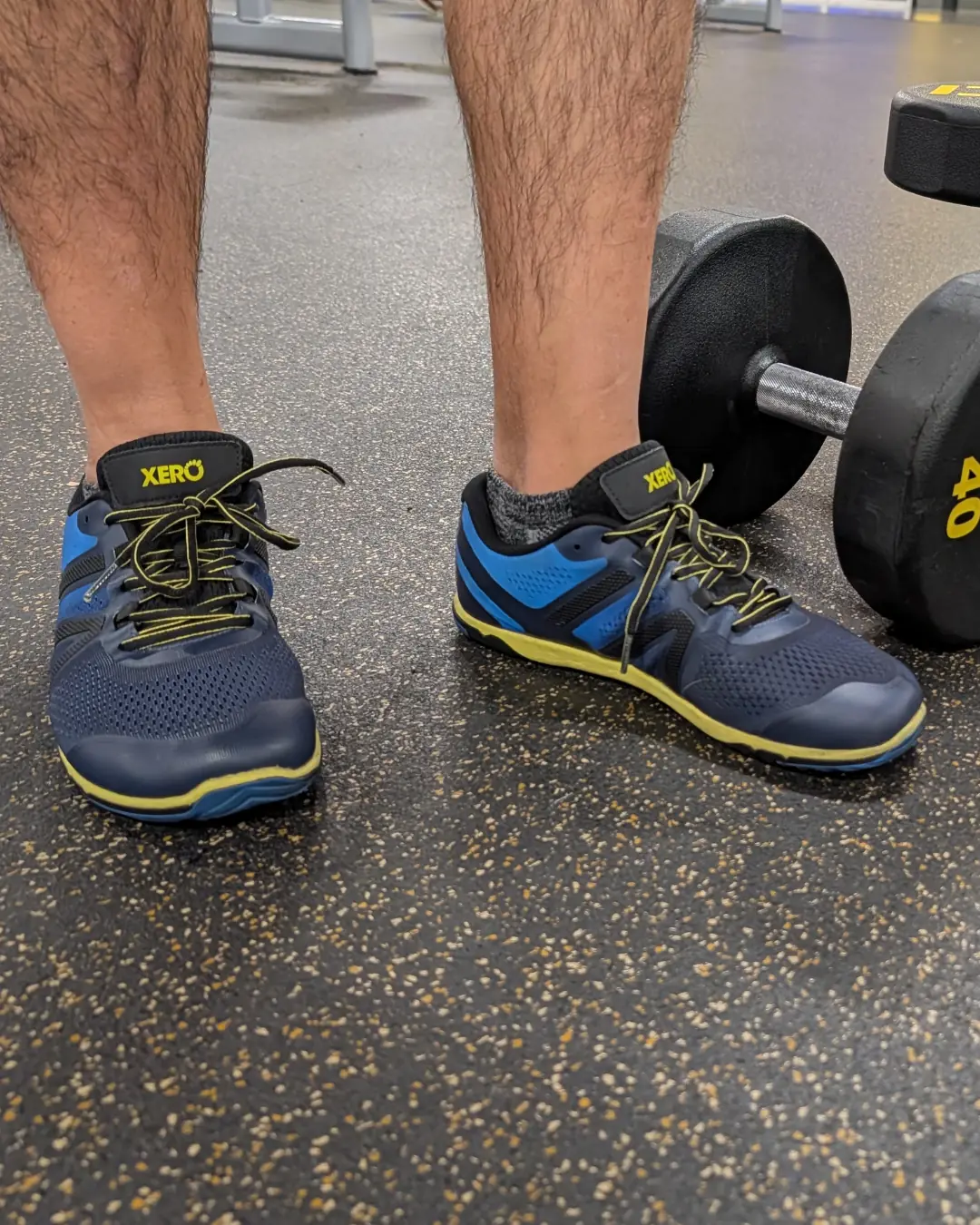 A person's lower legs and feet in navy and blue Xero Shoes minimalist trainers with thin yellow soles, standing beside a dumbbell on a gym floor